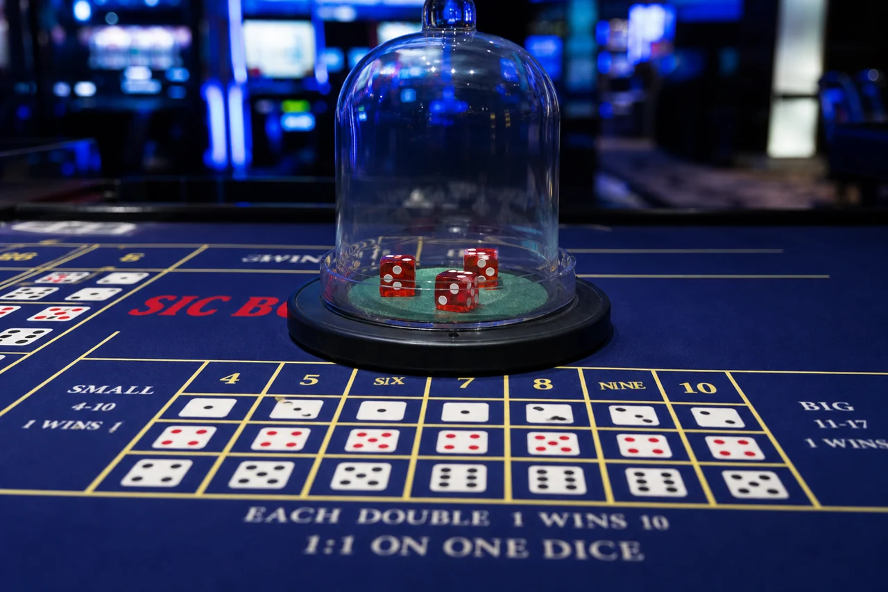Casino sic bo table with three red dice inside a clear bell shaker and visible betting grid, close realistic photograph, midnight blue casino lighting